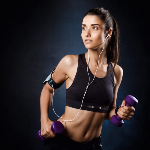 Young beautiful bruntete sportive girl training with dumbbells over dark background. Copy space.